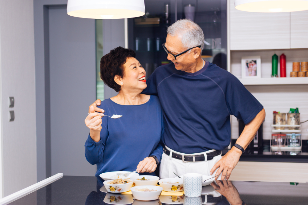 An elderly couple smiling at each other. There is food laid out on a table in front of them and the woman is holding a spoon of food